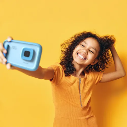 myFirst Camera 50 sininen lapsiystävällinen kamera, kuvassa lapsi ottaa selfien keltaista taustaa vasten